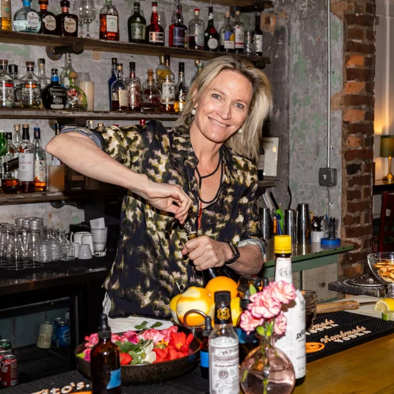A smiling bartender in a leopard print shirt prepares a drink at a rustic bar.