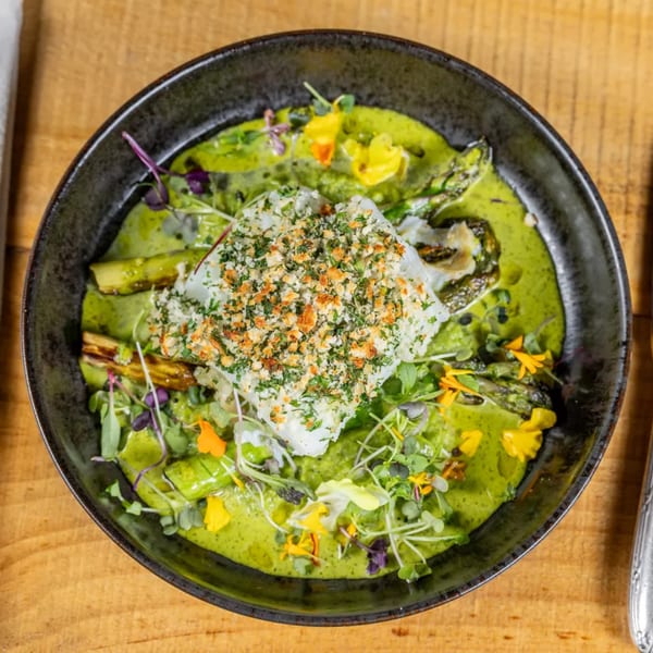 Cod with breadcrumbs and herbs, green sauce, asparagus, and edible flowers in a black bowl.