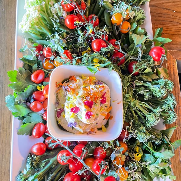 A white platter with a dip bowl surrounded by fresh vegetables like tomatoes, broccoli, and cauliflower.