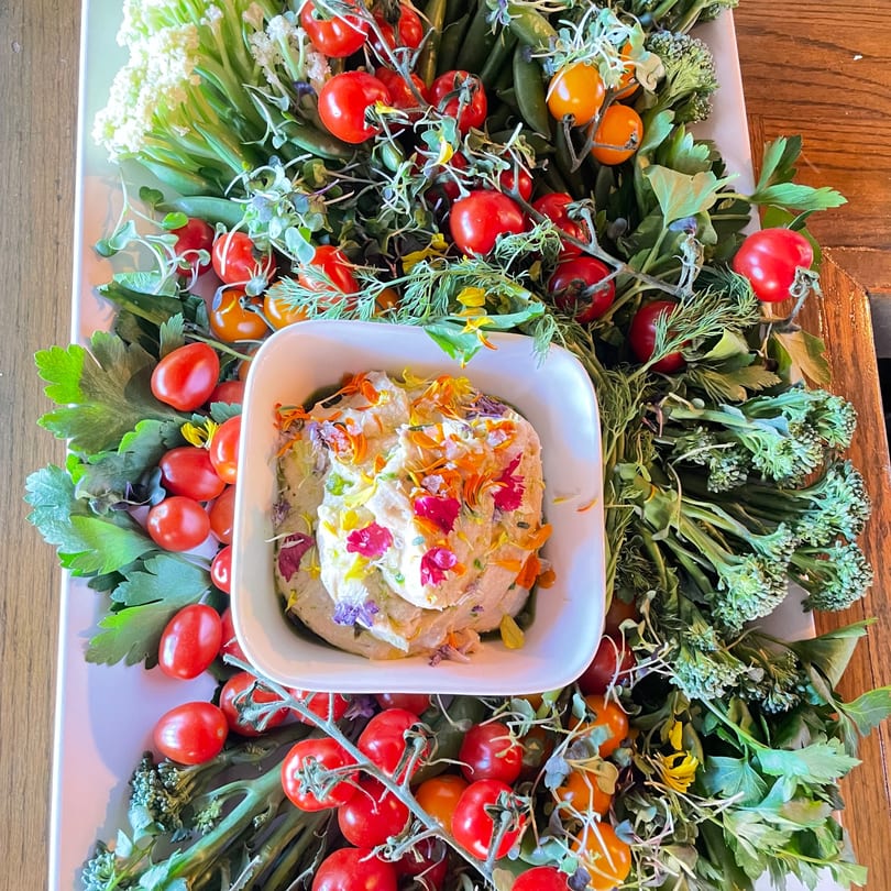 A white platter with a dip bowl surrounded by fresh vegetables like tomatoes, broccoli, and cauliflower.