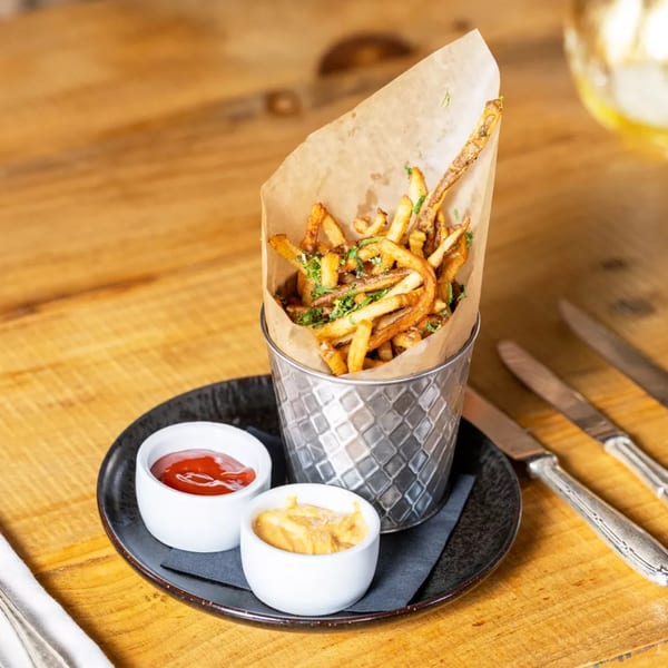 Crispy french fries in a metal bucket with ketchup and mustard on a wooden table.