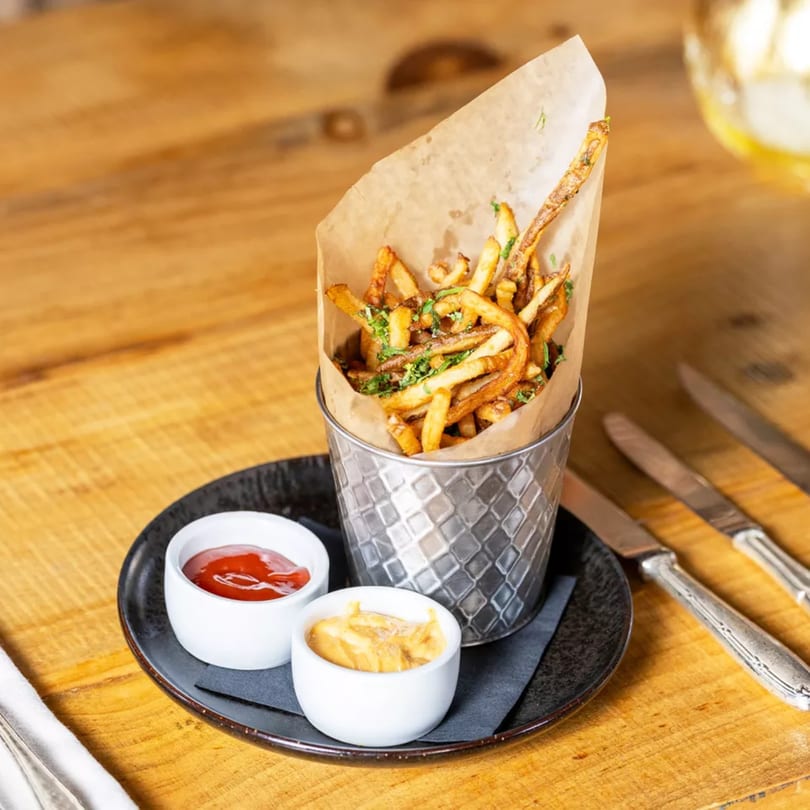 Crispy french fries in a metal bucket with ketchup and mustard on a wooden table.