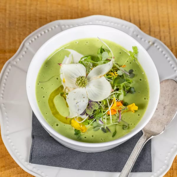 Vibrant green soup in a white bowl, garnished with microgreens, edible flowers, and olive oil.