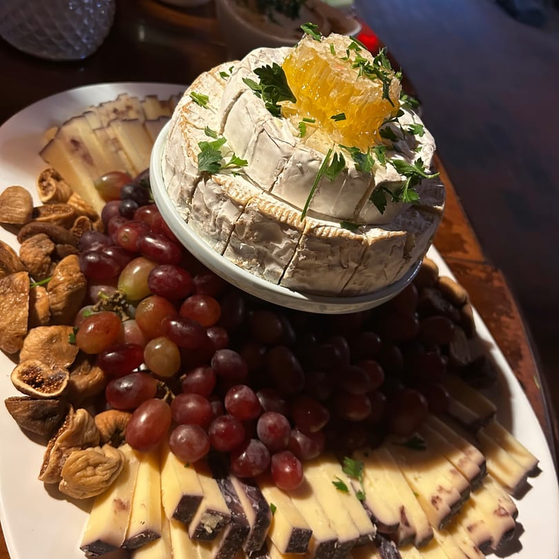 A cheese board with a wheel of brie topped with honeycomb, surrounded by grapes, figs, and sliced cheeses.