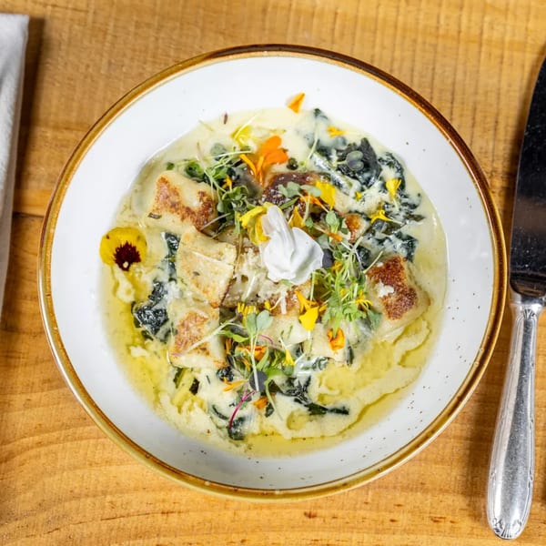 A white bowl of creamy gnocchi with spinach, microgreens, and edible flowers on a wooden table.