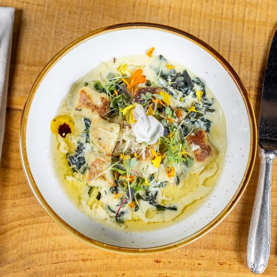 A white bowl of creamy gnocchi with spinach, microgreens, and edible flowers on a wooden table.