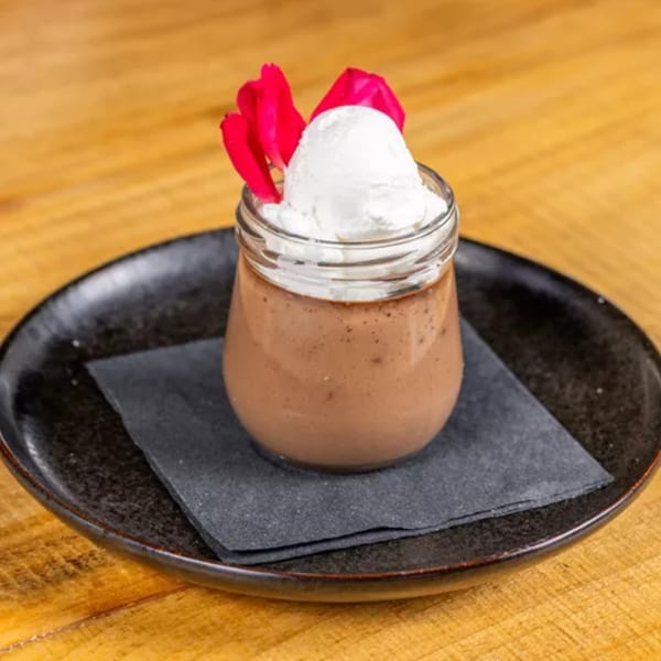 Chocolate pudding in a glass jar, topped with white cream and pink flower petals on a black plate.