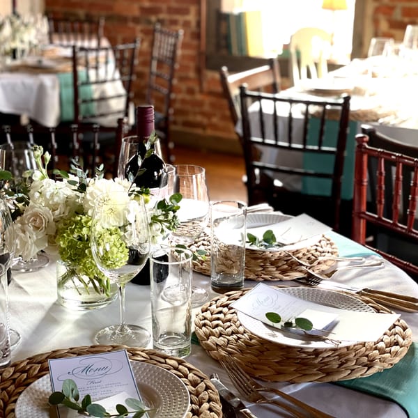 Elegantly set dining table with white tablecloths, woven placemats, wine, and floral centerpieces.