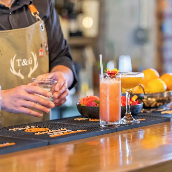 Bartender in apron preparing drinks on a wooden bar with two cocktails and citrus garnishes.