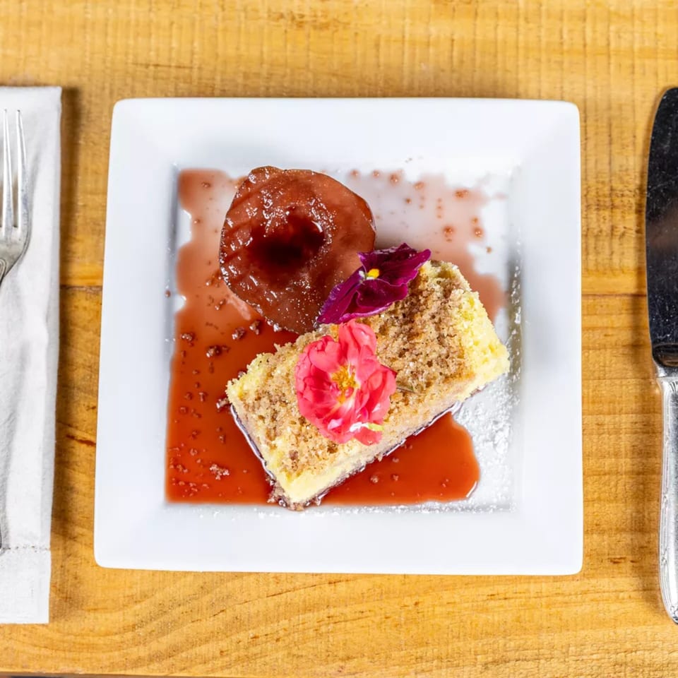 Cheesecake with crumble topping, poached pear, and edible flowers on a white plate.