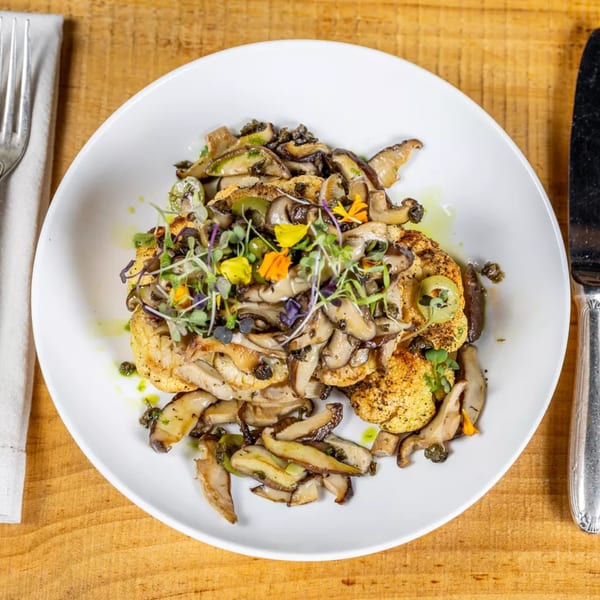 Pan-seared cauliflower steak topped with sautéed mushrooms, microgreens, and edible flowers on a white plate.