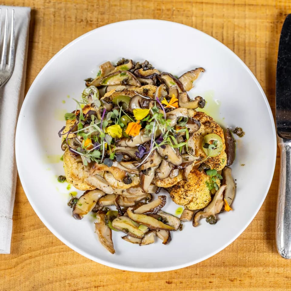 Pan-seared cauliflower steak topped with sautéed mushrooms, microgreens, and edible flowers on a white plate.