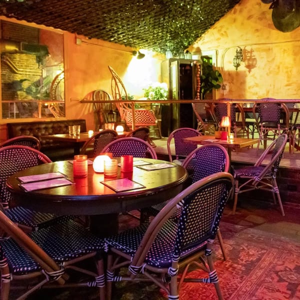 Cozy restaurant interior with round tables, wicker chairs, warm lighting, and a leafy ceiling.